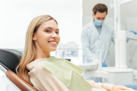 Foto de Joven hermosa mujer sonriente, paciente sentada en la clínica dental moderna con dentista en el fondo. Tratamiento dental, concepto de atención médica - Imagen libre de derechos