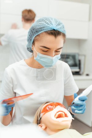 Téléchargez les photos : Dentiste féminine en uniforme de protection faisant le remplissage pour la patiente en clinique dentaire. Guérison des dents dans une clinique dentaire moderne - en image libre de droit