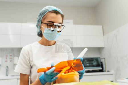 Foto de Retrato de un hermoso dentista en binoculares dentales examinando los dientes del paciente sentado en la silla en la odontología moderna. Tratamiento médico profesional femenino, limpieza. Concepto de cuidado dental - Imagen libre de derechos