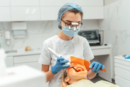 Téléchargez les photos : Patiente avec dentiste à la clinique. Médecin femme faisant procédure de blanchiment des dents avec la lumière ultraviolette lampe UV. Concept de blanchiment des dents - en image libre de droit