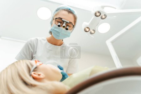 Téléchargez les photos : Portrait d'une dentiste attirante en jumelles dentaires examinant les dents d'une patiente assise dans une chaise en dentisterie moderne. Médecin professionnel féminin traitement, nettoyage. Concept de soins dentaires - en image libre de droit