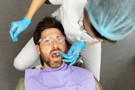 Photo for Portrait of handsome, middle aged patient sitting in dental chair wearing protective glasses with his mouth open. Dentist using tools to treat teeth. Oral hygiene, teeth treatment concept - Royalty Free Image