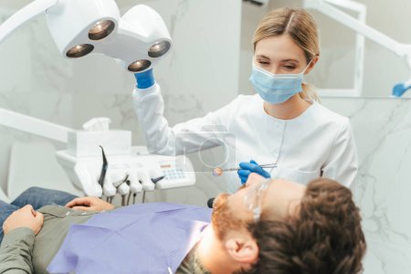 Photo for Professional woman dentist turning lamp while preparing checking man teeth. Young beautiful smiling female doctor holding mirror equipment in modern dental clinic. Teeth treatment, care concept - Royalty Free Image