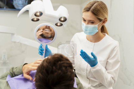 Photo for Portrait of dentist wearing protective mask, consultation patient. Male patient sitting in dental chair looking in mirror in modern clinic. Visit to doctor, treatment, teeth whitening - Royalty Free Image