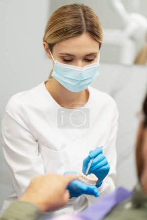 Téléchargez les photos : Dentiste blonde en gants chirurgicaux bleus, tenant un modèle de mâchoire humaine, tout en expliquant le traitement dentaire au patient masculin en clinique dentaire - en image libre de droit
