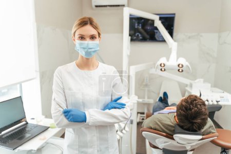 Téléchargez les photos : Dentiste féminine attrayante portant un masque de protection, debout dans un cabinet dentaire moderne, bras croisés, regardant la caméra. Patient masculin assis dans une chaise dentaire. Concept de traitement des dents - en image libre de droit