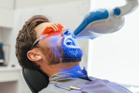Téléchargez les photos : Homme barbu beau patient blanchiment des dents dans la clinique dentaire moderne. Traitement des dents, concept de soins de santé - en image libre de droit