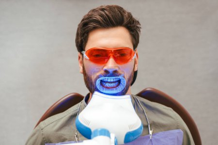 Photo for Portrait of handsome bearded middle aged man sitting in dental chair wearing red eyeglasses with open mouth, teeth whitening. Male patient in modern clean dentistry. Concept of dental treatment - Royalty Free Image