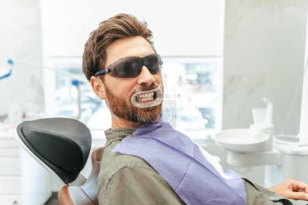 Téléchargez les photos : Beau client homme avec rétracteur à lèvres dans la bouche assis dans une chaise dentaire et regardant la caméra en attendant la procédure de blanchiment. Dents santé et dentisterie concept - en image libre de droit