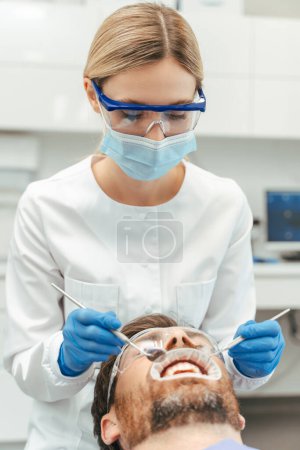 Photo for Blonde female dentist in latex gloves and uniform examining teeth of male patient laying in dental chair in modern clinic. Teeth health concept - Royalty Free Image