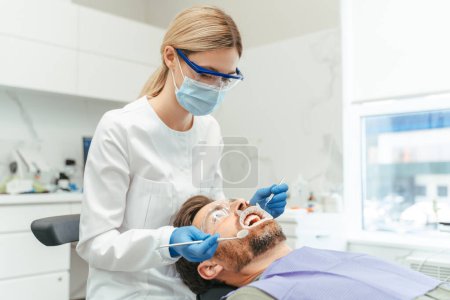 Téléchargez les photos : Portrait de beau dentiste avec des dents blondes de traitement des cheveux d'attrayant patient masculin barbu. Homme assis dans une chaise dentaire lors d'une visite à l'orthodontiste. Soins dentaires, soins de santé - en image libre de droit