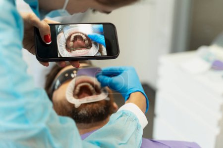 Foto de Paciente masculino con boca abierta sentado en silla dental, dentista sosteniendo teléfono móvil, tomando fotos. Concepto de tratamiento dental, cuidado dental, salud - Imagen libre de derechos