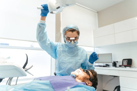 Téléchargez les photos : Chirurgien femelle avec microscope sur la tête faisant l'opération dentaire pour le patient masculin. Installation d'implants dentaires ou extraction dentaire à la clinique. Anesthésie générale en chirurgie orthodontique - en image libre de droit