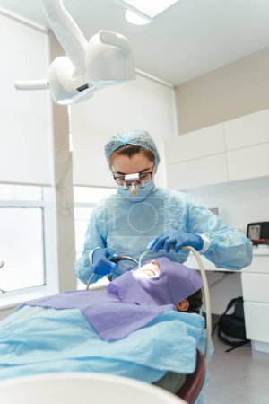 Photo for Portrait of attractive female dentist in surgical suit treatment patient's teeth in dental chair. Professional doctor in modern clinic using dental equipment and tools. Dental care concept - Royalty Free Image
