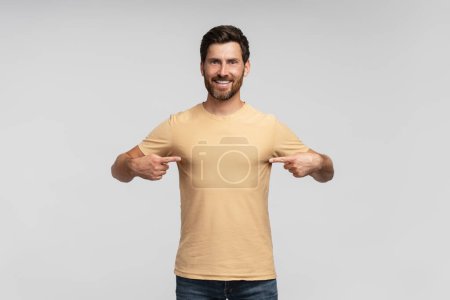 Téléchargez les photos : Homme barbu beau et souriant, hipster moderne portant un t-shirt blanc se pointant isolé sur fond gris. Maquette - en image libre de droit