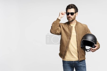 Téléchargez les photos : Homme barbu sérieux motard tenant casque de moto et regardant loin isolé sur fond gris. Modèle de mode veste en cuir marron, lunettes de soleil élégantes posant pour les photos - en image libre de droit