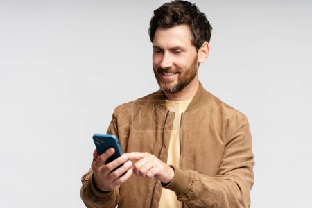 Foto de Retrato de hombre barbudo sonriente sosteniendo teléfono móvil, leyendo mensaje de texto, comunicación en línea aislada sobre fondo gris. Chico guapo de compras en línea, elegir en el sitio web - Imagen libre de derechos