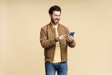 Téléchargez les photos : Portrait d'un homme barbu souriant tenant un téléphone portable, lisant un message texte, communiquant en ligne isolé sur fond beige. beau gars shopping en ligne, choisir sur le site - en image libre de droit