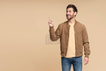 Téléchargez les photos : Un bel homme barbu souriant portant une veste d'automne marron isolée sur fond beige, pointant du doigt sur l'espace de copie. Shopping, vente, concept de magasin - en image libre de droit
