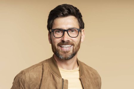 Téléchargez les photos : Portrait d'un bel homme barbu souriant portant des lunettes élégantes regardant la caméra isolée sur fond beige, espace de copie. Concept de vision - en image libre de droit