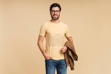 Foto de Guapo hombre barbudo sonriente sosteniendo chaqueta de otoño marrón mirando a la cámara aislada sobre fondo beige. Retrato de la exitosa modelo de mediana edad posando para fotos, toma de estudio - Imagen libre de derechos
