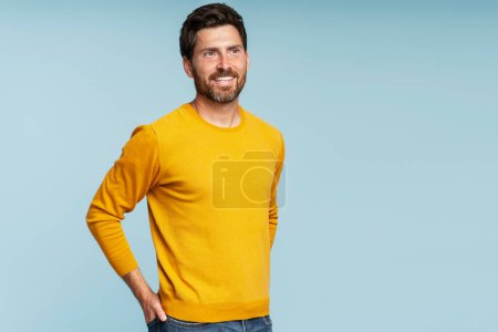 Foto de Retrato de un guapo hombre barbudo sonriente con suéter amarillo otoñal aislado sobre fondo azul. Retrato de la exitosa modelo de mediana edad posando para fotos, toma de estudio - Imagen libre de derechos