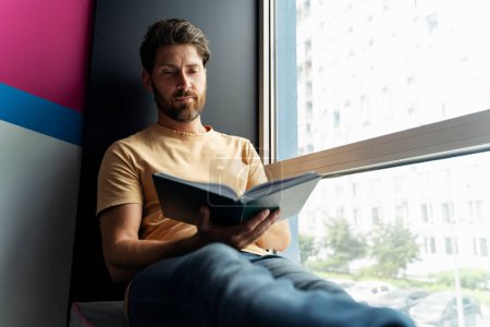 Photo for Portrait of handsome smiling man reading book sitting at home near window. Smart student studying, learning language, exam preparation in university campus. Education concept - Royalty Free Image