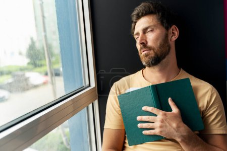 Foto de Hombre barbudo sobrecargado de trabajo durmiendo, sosteniendo el libro sentado cerca de la ventana. Retrato de estudiante universitario cansado en la biblioteca - Imagen libre de derechos