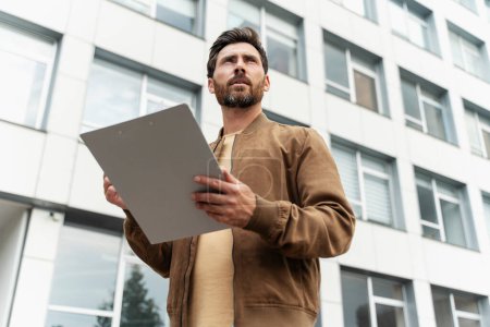 Photo for Confident bearded man holding business documents and looking away while standing on the street. Successful business, career concept - Royalty Free Image
