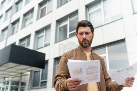 Photo for Handsome bearded businessman wearing stylish leather jacket looking at paper documents while standing near office building, copy space. Successful business concept - Royalty Free Image