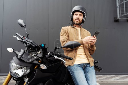 Téléchargez les photos : Homme élégant confiant, motard portant un casque à l'aide d'un téléphone portable debout près de moto sport. Beau modèle de mode portant une veste en cuir élégante regardant posant pour des photos sur moto - en image libre de droit