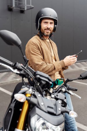 Téléchargez les photos : Homme élégant et confiant, motard portant un casque à l'aide d'un téléphone portable et regardant la caméra près de la moto sport. Beau modèle de mode portant une veste en cuir élégante posant pour des photos sur moto - en image libre de droit