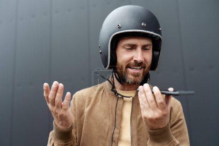 Photo for Stylish man biker wearing helmet recording voice message using mobile phone standing near sport motorcycle. Handsome fashion model wearing stylish leather jacket posing for pictures on motorbike - Royalty Free Image