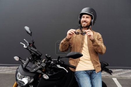 Téléchargez les photos : Homme de confiance souriant élégant, motard portant un casque debout près de la moto de sport. Beau modèle de mode réussi portant une veste en cuir élégante, des lunettes de soleil posant pour des photos sur moto - en image libre de droit
