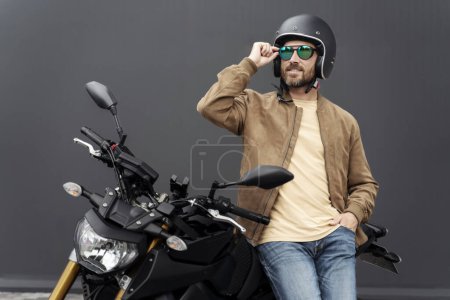 Téléchargez les photos : Brutal homme barbu, motard portant un casque debout près de moto de sport dans la rue. Beau modèle de mode réussi portant une veste en cuir élégante posant pour des photos sur moto - en image libre de droit