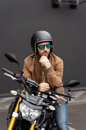 Téléchargez les photos : Homme barbu brutal, motard portant un casque assis à la moto de sport dans la rue. Beau modèle de mode réussi portant une veste en cuir élégante posant pour des photos sur moto - en image libre de droit