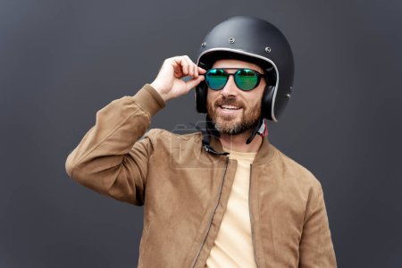 Foto de Motociclista barbudo feliz con casco de motocicleta y gafas de sol elegantes mirando hacia otro lado mientras se prepara paseo en moto. Aventura, libertad, concepto de viaje por carretera - Imagen libre de derechos