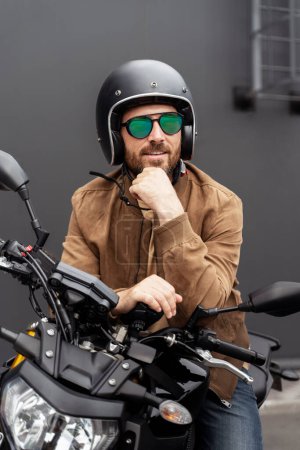 Téléchargez les photos : Portrait d'un bel homme barbu souriant, motard portant un casque, lunettes élégantes, veste en cuir posant sur la moto dans la rue. Voyage, liberté, concept de transport - en image libre de droit