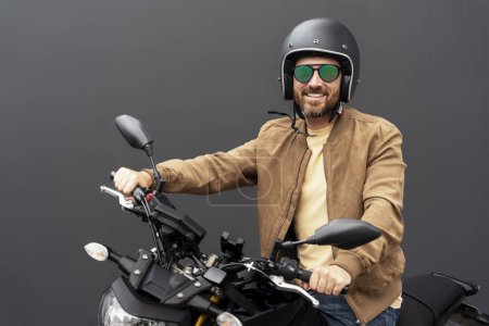 Téléchargez les photos : Portrait d'un bel homme barbu souriant, motard portant un casque, des lunettes élégantes, une veste en cuir à moto isolée sur fond noir. Voyage, liberté, concept de transport - en image libre de droit