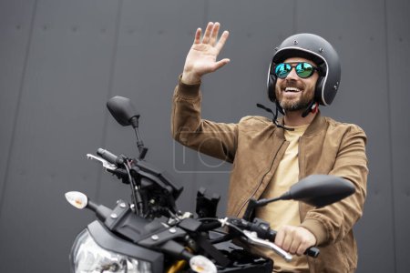 Téléchargez les photos : Joyeux motard barbu portant un casque et des lunettes de soleil élégantes agitant la main à quelqu'un assis sur la moto sur la route. Aventure, liberté, concept de road trip - en image libre de droit