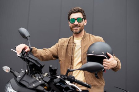 Photo for Stylish smiling confident man, biker holding helmet and looking at camera on sport motorcycle. Handsome successful fashion model wearing stylish leather jacket, sunglasses posing for pictures - Royalty Free Image