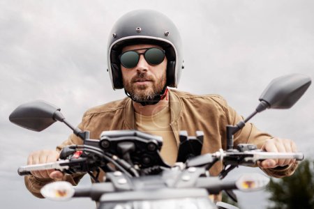 Téléchargez les photos : Beau motard barbu brutal portant un casque et des lunettes de soleil élégantes à moto sur la route. Aventure, liberté, concept de road trip - en image libre de droit