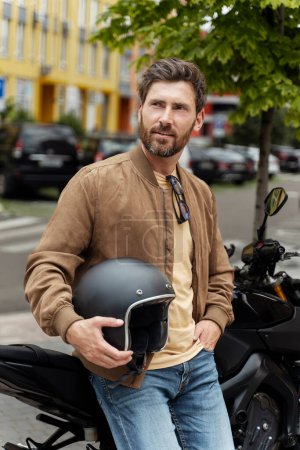 Photo for Stylish bearded brutal man, biker holding helmet standing near sport motorcycle on the street. Handsome successful fashion model wearing stylish leather jacket posing for pictures on motorbike - Royalty Free Image