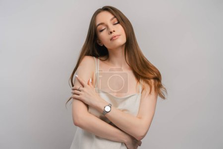 Photo for Portrait of cute woman hugging herself with watch on her hand isolated on gray background. Beautiful female wearing beautiful nightgown posing in studio with closed eyes. ?oncept of sexuality - Royalty Free Image