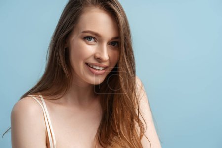 Téléchargez les photos : Portrait de beauté de jeune femme souriante aux cheveux longs regardant la caméra isolée sur fond bleu. Beauté naturelle, soin des cheveux, concept de routine matinale - en image libre de droit