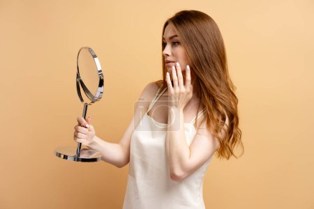 Téléchargez les photos : Belle femme sexy portant une nuisette touchant son visage en regardant dans un miroir isolé sur fond beige. routine du matin, concept de beauté naturelle - en image libre de droit