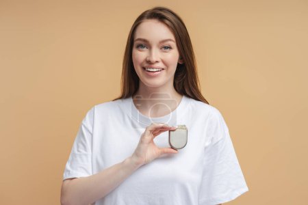 Photo for Young smiling woman holding cardio stimulator, pacemaker looking at camera isolated on beige background. Health care, cardiology, heart stimulation concept - Royalty Free Image