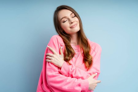 Photo for Portrait of cute woman hugging herself with closed eyes isolated on blue background. Beautiful female wearing pink sweatshirt posing in studio - Royalty Free Image