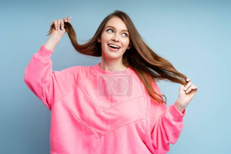Photo for Beautiful smiling caucasian woman wearing pink sweatshirt touching her shiny hair isolated on blue background. Attractive fashion model posing for picture in studio. Beauty, hair care concept - Royalty Free Image
