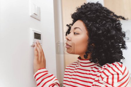 Foto de Retrato de un hermoso afroamericano serio parado en el apartamento usando el panel de control del hogar inteligente, ajustando la temperatura. Atractiva mujer. Concepto de casa inteligente - Imagen libre de derechos